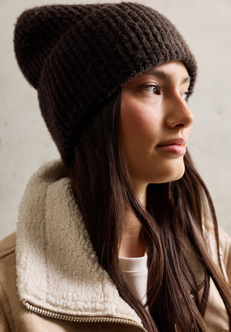 Braune Strickmütze mit einem strukturierten Muster, getragen zu einer beigen Jacke mit Lammfellkragen. Der Hintergrund ist neutral und unauffällig.