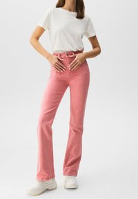 Jean flares en denim rose avec taille haute et poches avant, associés à un T-shirt blanc à manches courtes et des baskets blanches.