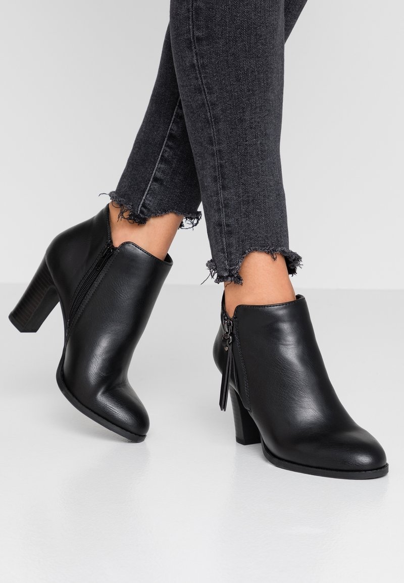 Botas de piel negra hasta el tobillo con punta redondeada, cierre de cremallera y un tacón cuadrado texturizado. Llevadas con jeans negros desgastados.