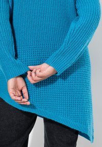 Pull en tricot turquoise avec une surface texturée et des poignets côtelés, doté d'un ourlet asymétrique et d'une coupe décontractée.