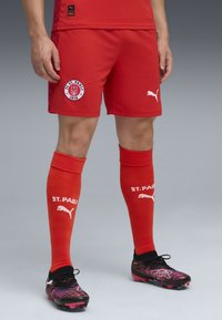Röda fotbollsshorts med en cirkulär FC St. Pauli-logotyp, matchande knästrumpor i röd färg med vit branding, och svarta fotbollsskor med livfulla mönster.