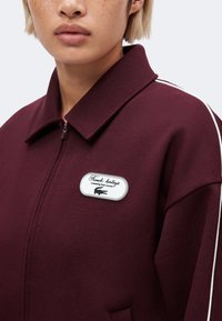 Femme portant une veste bordeaux zippée avec une étiquette blanche indiquant "French heritage Created by Rene Lacoste" et des bandes blanches sur les manches.