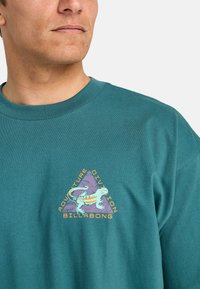 Teal cotton t-shirt featuring a graphic design with a triangle, lizard illustration, and text "Adventure Division Billabong" in contrasting colors.