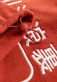 Piros polár anyag, zsinórral és kör alakú melltartóval, fehér hímzett szöveggel és mintákkal díszítve. Puha, texturált felület.