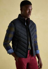 Mann in schwarzer Steppweste über einem hochgerollten karierten Hemd und roten Hosen, steht mit einer Hand in der Tasche vor einem beigen Hintergrund.