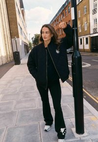 Pihe-puha fekete bomberdzseki arany cipzárral és csillag kitűzővel, fekete, bővülő szárú nadrággal, fehér grafikával. A fehér tornacipő teszi teljessé az összeállítást.