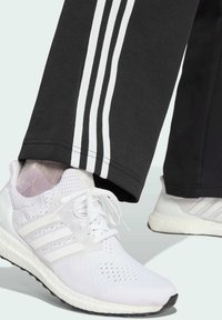 Weiße Sportschuhe mit Mesh-Obermaterial, drei gestreiften Akzenten und weißen Schnürsenkeln. Kombiniert mit schwarzen Hosen, die drei weiße vertikale Streifen haben.