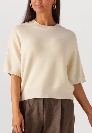 Frau trägt einen cremefarbenen Strickpullover mit kurzen Ärmeln, braune hochtaillierte gefaltete Hosen und goldene Creolen vor einem schlichten Hintergrund.