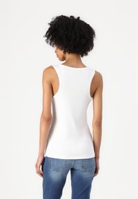 Weißes geripptes Tanktop mit breiten Trägern, das über einen runden Ausschnitt und eine figurbetonte Passform verfügt. Kombiniert mit hellblauen Jeans.
