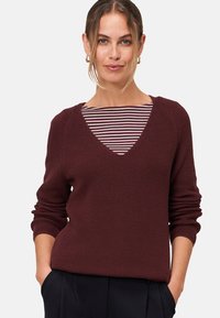 Burgundy knitted sweater with a V-neck and ribbed texture, layered over a striped shirt. Features long, slightly puffed sleeves.