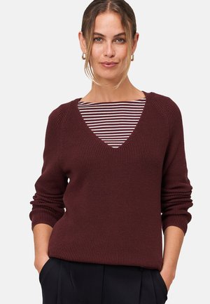 Pull en tricot bourgogne avec un col en V et une texture côtelée, superposé sur une chemise rayée. Présente des manches longues légèrement bouffantes.