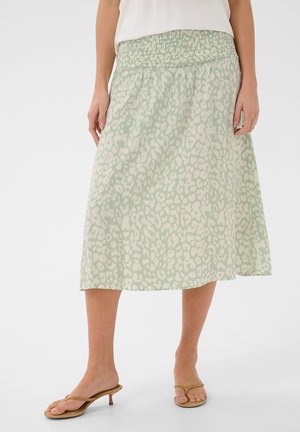 Žena nosí midi sukňu so zvieracím vzorom v svetlozelenej a béžovej farbe s širokým elastickým pásom a hnedými sandálmi na podpätku.