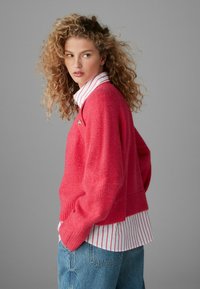 Lys pink strikket sweater med ribbede manchetter, lagdelt over en hvid og pink stribet skjorte med krave, parret med lyseblå denimjeans.