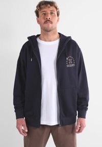 Sweat-shirt zippé bleu marine avec une poche kangourou, une capuche avec cordon de serrage et un motif graphique sur le devant. Porté par-dessus un t-shirt blanc à col ras du cou.