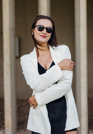 Bílý blazer přes černý top, doplněný tmavými slunečními brýlemi. Hladká látka s přiléhavým střihem a detailem s dvojitým knoflíkem.
