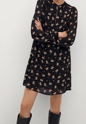 Schwarzes Blumenkleid mit langen Ärmeln, das einen dezenten Reißverschluss am Halsabschluss aufweist. Hergestellt aus leichtem Stoff, verziert mit kleinen, beigen Blumenmustern.