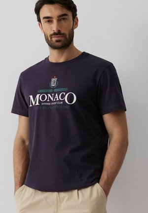 Brodaty mężczyzna o ciemnych włosach, ubrany w granatowy T-shirt z napisem "MONACO Diverse Sport Club" i małym emblematem, ręce w kieszeniach beżowych spodni.