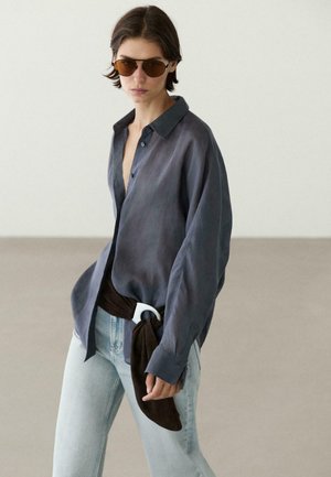 Osoba nosząca za dużą ciemnoszarą koszulę na guziki, jasnoniebieskie dżinsy, brązowy pasek-szalik z białą klamrą i brązowe okulary przeciwsłoneczne, stojąca w pomieszczeniu.