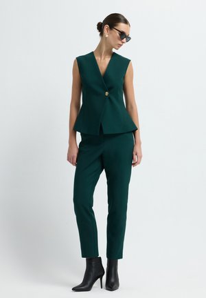 Gilet ajusté vert avec un décolleté en V et un bouton doré, associé à un pantalon slim et des bottines noires. Tissu lisse avec une forme structurée.