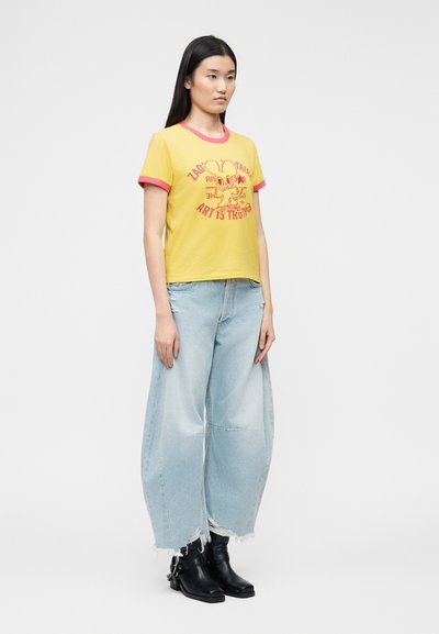 Γυναίκα με κίτρινο T-shirt με κόκκινα περιθώρια και τυπωμένο καρτούν, ανοιχτό μπλε τζιν παντελόνι με φαρδιά μπατζάκια και ξεφτισμένα τελειώματα, και μαύρα μποτάκια αστραγάλου.