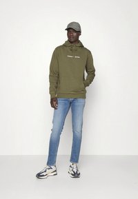 Olivzaļš džemperis ar kapuci, priekšējā kabata un logotips, gaiši zili plāni džinsi, pelēka cepure un daudzkrāsainas sporta kurpes ar teksturētu dizainu.