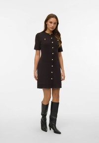 Vestito marrone in maglia con maniche corte, dettagli con bottoni dorati sulla parte frontale e una forma aderente. Indossato con stivali neri in pelle fino al ginocchio.