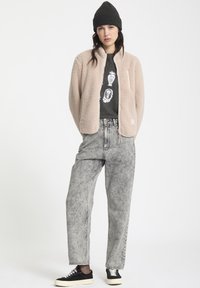 Lichtroze fleecejack met een ritssluiting aan de voorkant, zwart T-shirt met grafische print, grijze high-waisted jeans en zwarte sneakers met crèmekleurige zolen.