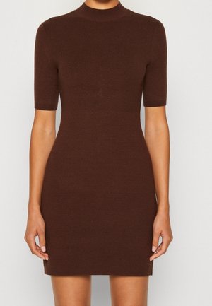 Kobieta ubrana w dopasowaną, krótką sukienkę dzianinową z krótkim rękawem i wysokim kołnierzem, stojąca na tle jednolitego jasnego tła.