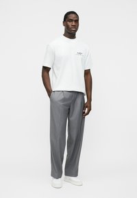 Mann iført hvit T-skjorte med grafisk trykk, grå plisserte bukser og hvite joggesko, stående med en hånd i lommen mot en ensfarget bakgrunn.