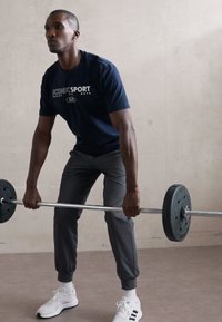 Atleta a levantar uma barra, vestindo uma camisa azul marinho com texto branco, calças cinza ajustadas e sapatos de desporto brancos com detalhes pretos.