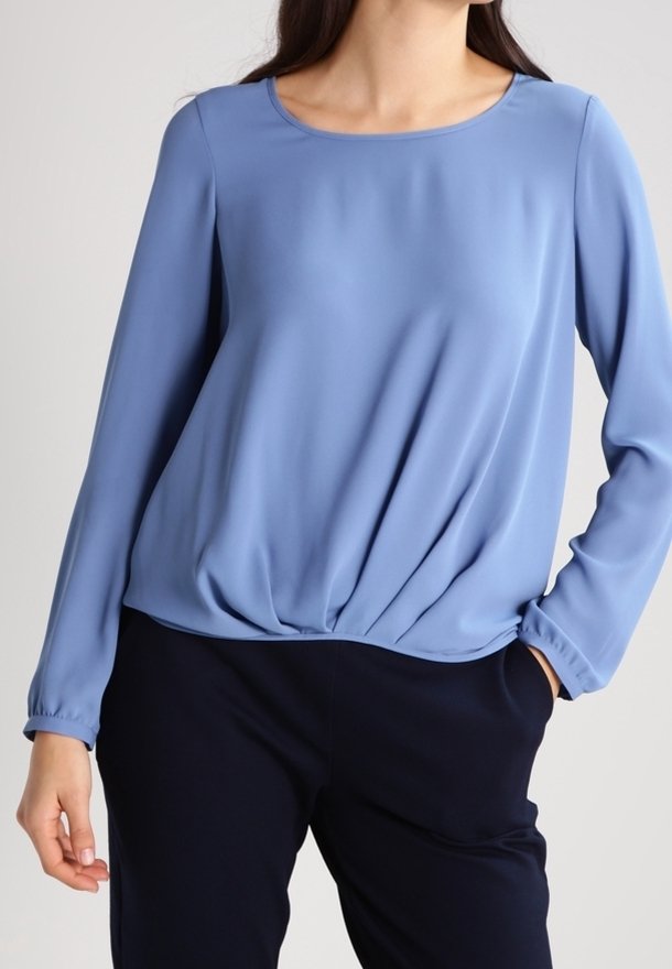 Femme portant un chemisier bleu doux à manches longues avec un ourlet froncé, associé à un pantalon noir, debout avec une main dans la poche.