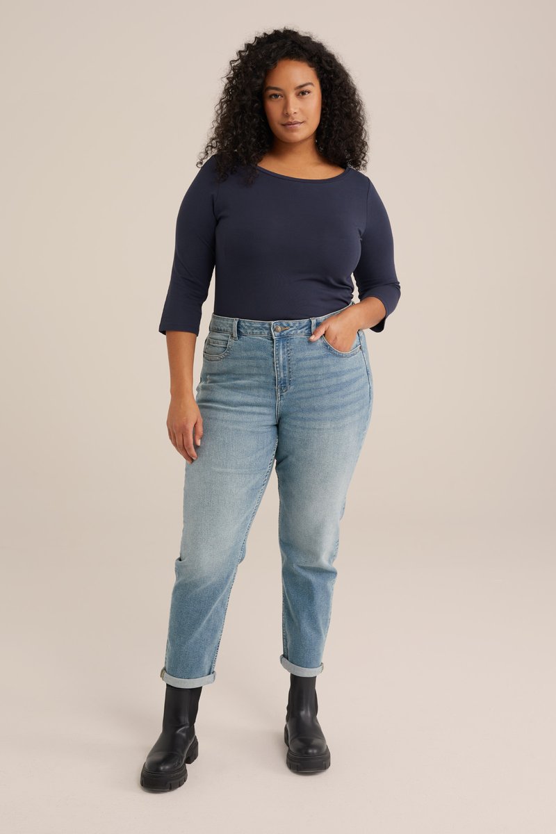 Kvinde med krøllet hår iført en marineblå top, lyseblå jeans med opkrogede kanter og sorte ankelstøvler, stående mod en ensfarvet baggrund.
