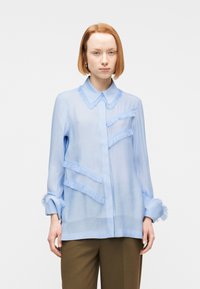 Νεαρό άτομο με κοντά κόκκινα μαλλιά, φορώντας ανοιχτό μπλε μπλούζα με φούντες και καφέ παντελόνι, πάνω σε απλό  λευκό φόντο.