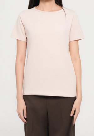 Femme portant un t-shirt à manches courtes beige clair uni et un pantalon brun foncé, debout avec les bras détendus le long du corps devant un fond blanc.