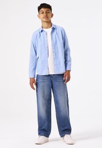 Camisa de botones a rayas azul claro sobre una camiseta blanca, combinada con vaqueros azules holgados y zapatillas blancas. Diseño simple, ajuste informal.