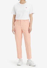 Vit t-shirt med en liten röd logotyp, kombinerad med ljusrosa byxor och vita sneakers. Outfiten har en avslappnad passform och rena linjer.