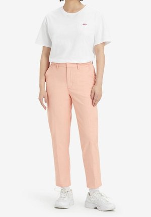 T-shirt blanc avec un petit logo rouge, associé à un pantalon rose clair et des baskets blanches. La tenue présente une coupe décontractée et des lignes épurées.
