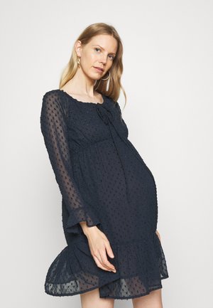 Vestido de maternidad azul marino con patrón de puntos texturizados, dobladillo con volantes, mangas largas translúcidas y detalle de lazo en el escote.
