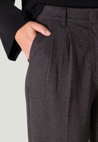 Dunkelgraue, maßgeschneiderte Hose aus strukturiertem Stoff, mit klassischer Falte und Seitentaschen. Nahaufnahme mit Hand in der Tasche.