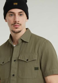Olijfgroen overhemd met korte mouwen, twee borstzakken, zwarte knopen en een labelpatch. Draagt een zwarte gebreide beanie.