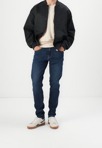 Chaqueta bomber negra, camisa beige, pantalones vaqueros azules con un ligero desgaste y zapatillas blancas con acentos negros. Manos en los bolsillos, pose de pie.