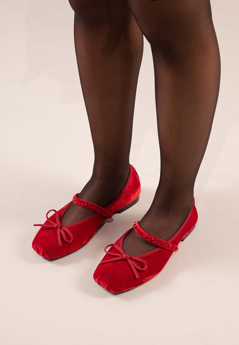 Bailarinas de terciopelo rojo con punta cuadrada, un lazo decorativo y una correa adornada con pequeños tachuelas. Llevadas con medias negras.
