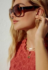 Överdimensionerade sköldpadds solglasögon med gradientbruna linser. Modellen bär en texturerad koralltop och lager av guldfärgade halsband.