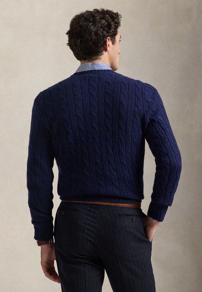 Μπλε ναυτικό πλεκτό πουλόβερ με σχέδιο καλώδιου, ριμπ μανσέτες και τελείωμα. Φορεμένο πάνω από ένα ανοιχτό μπλε και λευκό ριγέ πουκάμισο. Συνδυασμένο με παντελόνια με λεπτές ρίγες.