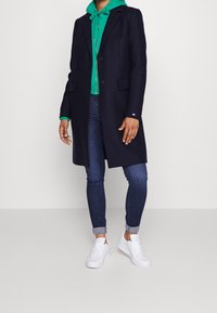 Cappotto in misto lana blu navy con due bottoni, abbinato a una felpa verde con cappuccio e jeans arrotolati. Sneakers bianche completano l'outfit.