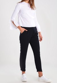 Witte longsleeve top met gestrikte accenten bij de mouwen, gecombineerd met zwarte tapse broek en witte sneakers, tegen een neutrale achtergrond.