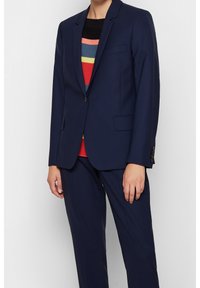 Mörkblå kostym med skräddarsydd passform, med en knappstängning, skärpkragar och sidofickor. Bärs över en flerfärgad randig tröja.