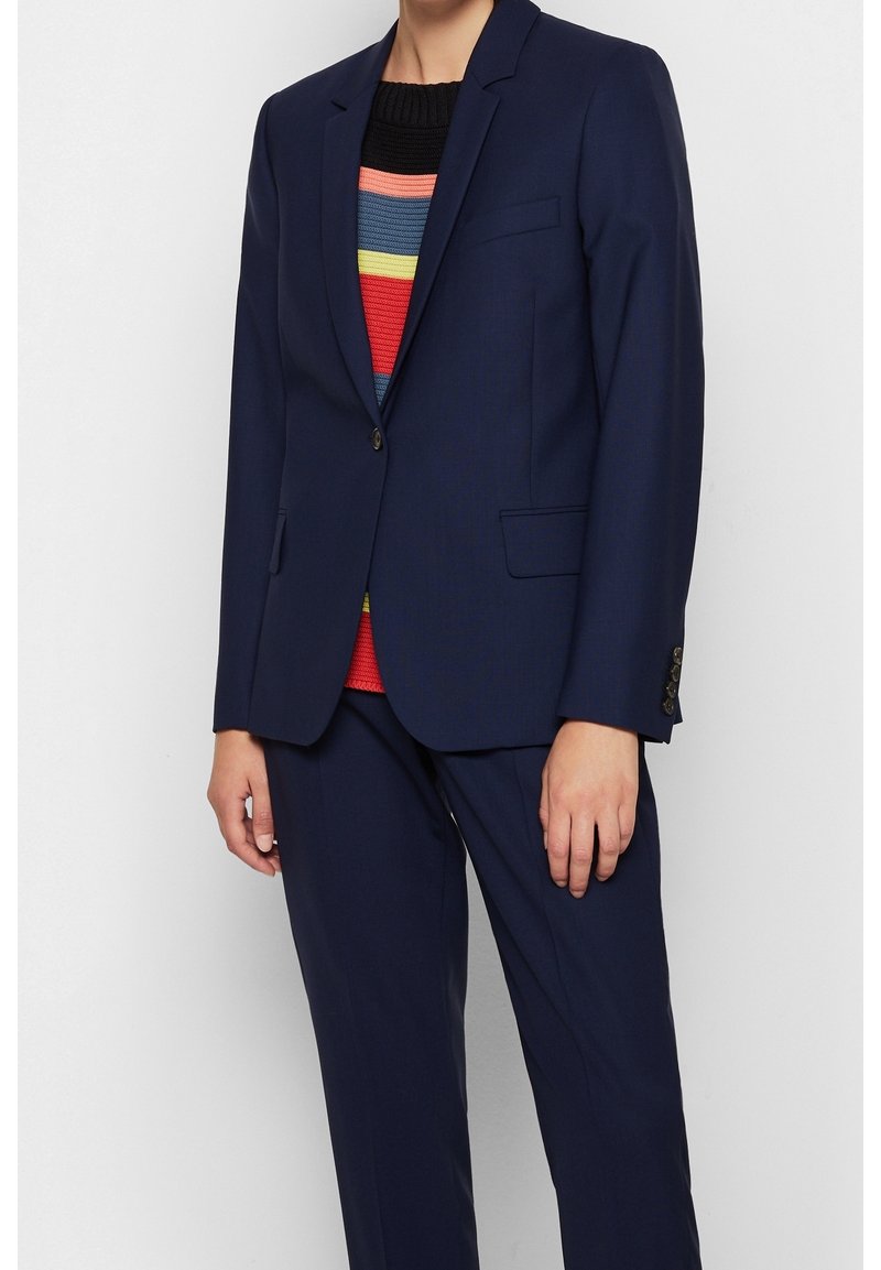 Mörkblå kostym med skräddarsydd passform, med en knappstängning, skärpkragar och sidofickor. Bärs över en flerfärgad randig tröja.