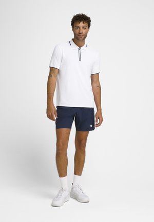 Mand iført en hvid kortærmet polo med marineblå shorts, hvide sokker og hvide sneakers, stående mod en ensfarvet hvid baggrund.