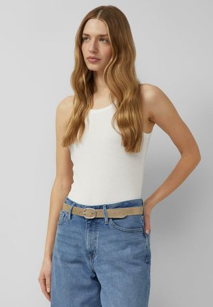 Kobieta z długimi falującymi włosami ubrana w biały bezrękawnik, niebieskie dżinsy i beżowy pasek, stojąca na tle jednolitego szarego tła.